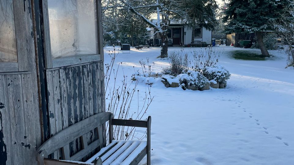 Verschneiter Gartenhof mit einer verwitterten Holzhütte und einer schneebedeckten Bank im Vordergrund. Im Hintergrund stehen große Nadel- und Laubbäume sowie ein kleines Wohnhaus, umgeben von Schnee und einigen Sträuchern.