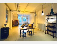 Industrieartiger Raum mit heller, minimal gestalteter Einrichtung; im Vordergrund ein Holztisch mit Hockern, im Hintergrund eine Sitzecke mit gelben Sofas und großen Fenstern. An den Wänden sind Industrielampen, ein Regal und ein Heizkörper sichtbar.