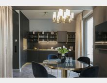 Moderne Küche mit dunklen Schränken, offenen Regalen und goldenen Akzenten, daneben ein runder Esstisch mit Glasplatte und blauen Stühlen. Im Vordergrund steht eine Vase mit weißen Tulpen, und an der Decke hängt ein eleganter Kronleuchter.