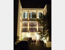 Nachtszene eines beleuchteten, mehrstöckigen Gebäudes mit verzierten Fassaden und großen Fenstern. Im Vordergrund stehen mehrere geparkte Fahrzeuge und eine Gruppe von Menschen, die sich vor dem Eingang des Gebäudes versammelt hat.