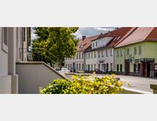 Straßenszene in einer kleinen Stadt mit Wohn- und Geschäftshäusern, die stilistisch schlicht gestaltet sind und rote Ziegeldächer haben. Ein Treppengeländer ist im Vordergrund sichtbar und Bäume säumen den Gehweg entlang der Straße.