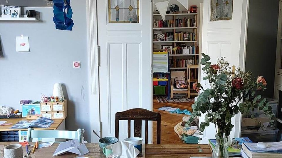 A cozy interior showing a cluttered wooden dining table with chairs, surrounded by shelves and cabinets containing books and small items. Double white doors with decorative stained glass panels lead to a room with more bookshelves and toys, highlighting a lived-in and creative atmosphere.