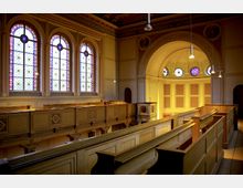 Innenraum einer historischen Synagoge mit verzierten Holzgalerien, farbigen Buntglasfenstern und gewölbter Decke. Links befinden sich drei große Fenster, die reich mit Blumen- und geometrischen Mustern gestaltet sind; rechts ist eine halbrunde Nische mit kleineren dekorativen Fenstern zu sehen.