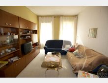 Wohnzimmer mit einer gelben Wand und zwei großen Fenstern mit weißen Vorhängen. Links steht eine Holz-Schrankwand mit Fernseher, rechts befinden sich ein dunkler Sofasessel und ein helles Sofa mit Decke und Kissen. In der Mitte steht ein Couchtisch aus Glas mit Dekorationen.