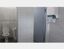 Öffentliche Toilettenanlage mit einer weißen Toilette, einem Handtrockner und einem Papierhandtuchspender. Die Wände sind mit hellen Fliesen verkleidet, der Boden ist dunkel gefliest, und eine graue Tür führt zu einem weiteren Bereich.