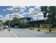 Eine erhöhte S-Bahn-Station mit einer verglasten Überdachung steht über einer Straße. Im Vordergrund sind Bürgersteige, ein Fahrradweg, parkende Fahrzeuge und Passanten zu sehen, umgeben von Bäumen und grüner Vegetation.