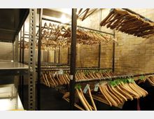 Raum mit Backsteinwänden und mehreren Metallstangen, an denen zahlreiche leere Holzbügel hängen; beleuchtet durch Deckenlampen. Im Vordergrund ist ein Regal sichtbar, im Hintergrund stehen einzelne Stühle.