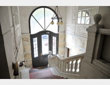 Treppenaufgang in einem historischen Gebäude mit Marmorelementen an den Wänden und einem geschwungenen Handlauf. Im Hintergrund eine große halbkreisförmige Glastür mit schwarzen Rahmen und darüber ein Kronleuchter mit drei Glühbirnen.