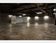 Große, leere Industriehalle mit Betonboden und dunklen Ziegelwänden, in der Mitte stehen mehrere freistehende weiße Stellwände. An der offenen Decke sind sichtbare Rohrleitungen, Stahlträger und Deckenleuchten angebracht.