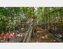 Kletterwald in einem dicht bewaldeten Gebiet mit verschiedenen Seilbrücken, Plattformen und Netzen in verschiedenen Höhen. Menschen mit Kletterausrüstung bewegen sich auf den Hindernissen, während am Boden andere Besucher und Begleitpersonen auf Holzwegen stehen.