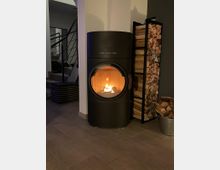 Moderner Innenraum mit einem schwarzen, runden Kaminofen der Marke "Austroflamm", in dem ein Feuer brennt. Daneben steht ein Regal, das mit gestapeltem Brennholz gefüllt ist, und ein Eimer mit weiteren Holzstücken steht auf dem Boden.