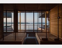 Innenraum einer modernen Sauna mit Holzverkleidung, großflächigen Fensterfronten und Blick auf einen ruhigen See. Draußen sind ein Holzsteg und Sitzgelegenheiten sichtbar, eingerahmt von minimalistischer Architektur.