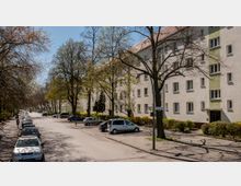 Mehrstöckige Wohngebäude mit heller Fassade und grünen Akzentfarben säumen eine baumbestandene Straße. Am Straßenrand parken Autos, und vereinzelte Fußgänger sind auf dem Gehweg unterwegs.