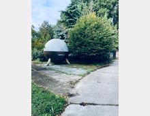 Kleiner, kugelförmiger Pavillon mit Metall- und Holzelementen, umgeben von dichten Bäumen und Büschen in einer parkähnlichen Umgebung. Ein gepflasterter Weg führt an der Konstruktion vorbei.