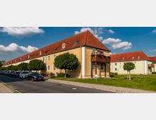 Reihenhaus in einer Wohnsiedlung mit gelben und hellgrünen Fassaden, roten Ziegeldächern und kleinen Dachfenstern. Vor den Gebäuden sind gepflegte Bäume, parkende Autos und ein Gehweg entlang einer asphaltierten Straße zu sehen.