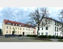 Mehrfamilienhäuser in einer Wohngegend mit hellgelben und weißen Fassaden, roten Ziegeldächern und modernen Fenstern. Im Vordergrund stehen Bäume, ein gepflasterter Platz und geparkte Autos.