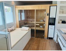Ein moderner Wellness-Raum mit einer Holzsauna mit Glastür, daneben ein Ruhebereich mit gepolsterter Sitzfläche und Kissen. Der Boden ist aus Holz, und es gibt ein Waschbecken sowie Regale mit Handtüchern und Pflegeutensilien.