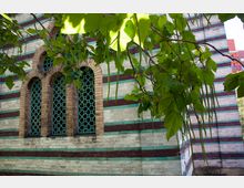 Fassade eines Gebäudes mit gestreiftem Mauerwerk aus abwechselnd hellen und dunklen Ziegelsteinen sowie grünen dekorativen Fenstern mit Gittermustern. Im Vordergrund hängen Zweige mit grünen Blättern von einem Baum herab.