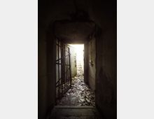Ein schmaler, dunkler Flur mit abblätternden Wänden und einer offenen Metallgittertür sowie einer schwer beschädigten Holztür. Der Boden ist mit Schutt und Laub bedeckt, während am Ende des Flurs Tageslicht sichtbar wird.