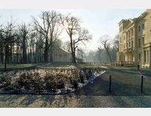 Ein historisches Gebäude mit hellgelber Fassade steht rechts im Bild, umgeben von einem gepflasterten Weg und winterlicher Vegetation. Im Hintergrund ist ein kleiner klassizistischer Pavillon inmitten eines Parks mit kahlen Bäumen zu sehen, durch die die Sonne scheint.