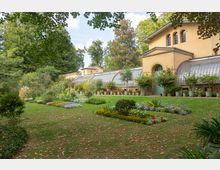 Gelber historischer Gewächshausbau mit großen Glasfenstern und Rundbogen-Türen, umgeben von gepflegten Beeten mit bunten Blumen und Pflanzen in Töpfen auf einem grünen Rasen, eingerahmt von Bäumen im Hintergrund.