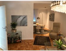 Wohnzimmer mit hellen Wänden und Parkettboden. Im Vordergrund ein Sessel, ein Glastisch und eine Zimmerpflanze; im Hintergrund ein runder Esstisch mit vier weißen Stühlen, einem Teppich und einer Bogenlampe. Links steht ein Barwagen mit Flaschen und Dekoration.