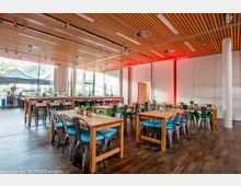 Ein moderner Veranstaltungsraum mit Holzboden, Holzdeckenpanelen und großen Fenstern, die viel Tageslicht hereinlassen. Der Raum ist mit Holztischen und Stühlen mit türkisfarbenen Sitzpolstern ausgestattet, und im Hintergrund sind Zelte und Grünflächen durch die Fenster sichtbar.