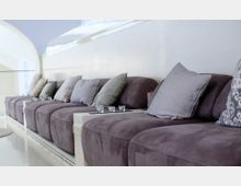 Lange Sitzbank mit violetten Polstern und verschiedenfarbigen Kissen vor einer weißen Wand, ergänzt durch kleine Teelichthalter auf weißen Zwischenablagen. Im Hintergrund sind helle, gewölbte Fenster- und Wandstrukturen sichtbar.