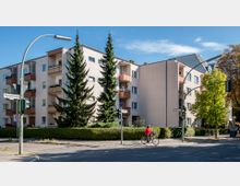 Ecke einer Wohnstraße mit einem mehrstöckigen, beige-weißen Wohnhaus mit Balkonen, umgeben von Bäumen, Büschen und Straßenschildern. Im Vordergrund fährt eine Person auf einem Fahrrad an einer Ampel vorbei.