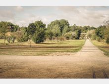 Weite Parklandschaft mit einem gepflasterten Weg, der durch eine Wiese führt, umgeben von Bäumen und Sträuchern. Im Vordergrund verlaufen Bahngleise, und im Hintergrund sind ein Aussichtsturm und ein Gebäude zu sehen.
