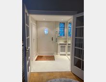 Eingangsbereich eines Wohnhauses mit einer weißen Tür, die ein ovales Fenster mit buntem Glas enthält. Rechts befinden sich blaue Glasfenster und eine weiße Heizkörperverkleidung, links zwei Türen; der Boden ist mit hellem Teppich und Parkett ausgelegt.