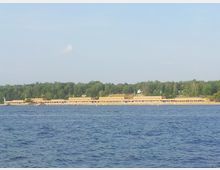 Ein großes Gebäude im Stil der klassischen Moderne erstreckt sich parallel zu einem Seeufer. Im Hintergrund ist ein dichter Wald zu sehen, während das Wasser im Vordergrund leicht wellig ist.