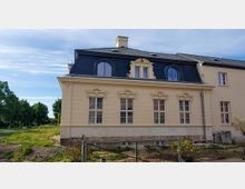 Beige-farbenes, zweistöckiges Gebäude mit Mansardendach und Dachgauben, darunter symmetrisch angeordnete Fenster mit ornamentalen Verzierungen. Der Vordergrund zeigt unbepflanzten Boden und einen Zaun, im Hintergrund sind Bäume und ein blauer Himmel sichtbar.