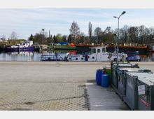 Hafenbereich mit mehreren Booten und Schiffen, die an einem ruhigen Gewässer liegen, umgeben von Bäumen und Gewerbebauten im Hintergrund. Im Vordergrund befinden sich Müllcontainer und blaue Kunststofftonnen auf einer betonierten Fläche.