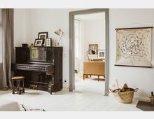 Wohnzimmer mit einem schwarzen Klavier und einem Hocker im Vordergrund, an einer Wand ein historisches Wetterkarten-Poster. Durch eine geöffnete Tür ist ein angrenzender Raum mit einem beige-farbenen Sofa und gerahmten Porträts an der Wand zu sehen. Ein Korb mit Holzscheiten steht rechts neben der Tür.