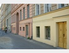 Straßenszene mit einer Reihe von Altbauwohnhäusern in Pastellfarben (rosa, beige und gelb). Auf dem gepflasterten Gehweg fährt eine Person mit einem Fahrrad, das Gebäude auf der rechten Seite hat vergitterte Fenster und eine gelbe Tür.