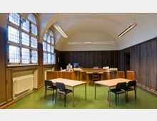 Innenraum eines Gerichtssaals mit hohen, gewölbten Decken, bleiverglasten Fenstern und Wandvertäfelung aus dunklem Holz. Im Vordergrund stehen zwei einfache Tische mit Stühlen, im Hintergrund ein erhöhtes Richterpult mit Computerbildschirmen.