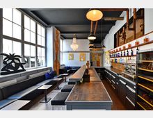 Moderner Barbereich mit einer langen Holztheke, schwarzen Sitzbänken und kleinen Beistelltischen. Die Wände sind weiß, teilweise mit freigelegtem Mauerwerk, Kunstwerken und dekorativen Elementen wie einer auffälligen Hängelampe sowie industriellen Rohrleitungen ausgestattet.