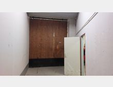 Innenansicht eines schlichten Raumes mit weißen Wänden und grauem Boden. Im Hintergrund befindet sich ein großes Tor aus Holz, daneben eine geöffnete Tür, die in einen anderen Bereich führt, in dem ein Feuerlöscher sichtbar ist.