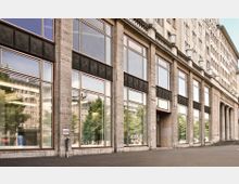 Facade of a large stone building with tall rectangular panes of reflective glass windows and decorative metal panels above them. The structure features classical architectural elements, such as columns and a balustrade, and is situated along a paved walkway.