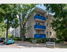 Wohngebäude in einer ruhigen Wohnstraße mit drei Stockwerken und blauen Balkonen, umgeben von großen Bäumen und Grünflächen. Im Vordergrund parken Autos am Straßenrand, und ein Gebäudeschild mit Hausnummern ist sichtbar.