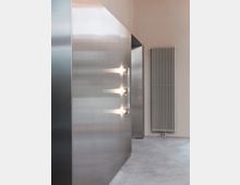 Minimalistischer Innenraum mit Wänden aus gebürstetem Metall, die durch runde, integrierte Leuchten beleuchtet werden. Rechts befindet sich ein weißer Heizkörper vor einer schlichten, beigefarbenen Wand, der Boden besteht aus poliertem Beton.