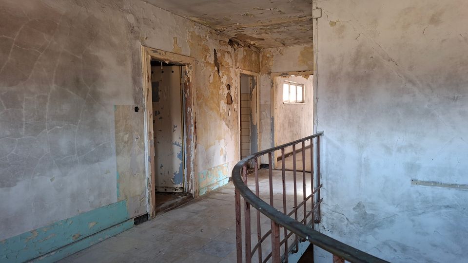 Verlassener Innenflur in einem stark renovierungsbedürftigen Gebäude mit abblätternder Farbe, rissigem Putz und mehreren Türöffnungen. Ein gebogenes Metallgeländer führt entlang eines Treppenabsatzes zu einem kleinen Fenster am Ende des Gangs.