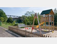 Spielplatz mit einem Holzklettergerüst und einer Rutsche, umgeben von einem niedrigen Holzzaun. Im Hintergrund fließt ein Fluss, dahinter Bäume und eine metallene Eisenbahnbrücke.