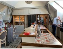 Ein geräumiger, rustikaler Dachküchenraum mit Holzdeckenbalken, einer Kücheninsel mit bunten Fliesen, und eingebauten Schränken in hellem Grau. Im Hintergrund ist ein Essbereich mit dekoriertem Tisch sowie ein Mann an der Kaffeemaschine zu sehen.