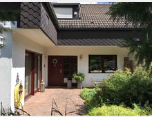 Eingangsbereich eines Einfamilienhauses mit heller Fassade, brauner Holztür, bodentiefen Fenstern mit Holzrahmen und einem Vorgarten mit vielen Pflanzen. Der gepflasterte Weg führt zur Haustür, und das Dach ist mit dunklen Ziegeln gedeckt.