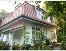 Zweistöckiges Wohnhaus mit weißer Fassade, roten Dachziegeln und mehreren Fenstern mit Fensterläden. Das Gebäude ist von Pflanzen und Bäumen umgeben und hat ein kleines Vordach sowie einen geschotterten Weg.