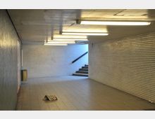 Unterführung mit weißen Fliesenwänden, einem Boden aus beigen Fliesen und Deckenbeleuchtung durch langgezogene Lampen. Im Hintergrund führen Treppenstufen nach oben, links ist ein Mülleimer an der Wand befestigt, und eine zerknüllte Zeitung liegt auf dem Boden.