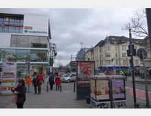 Straßenszene in einer belebten Stadt mit einem modernen Gebäude mit Glasfassade, das die Logos "DAK" und "Globetrotter" trägt, sowie historischen Gebäuden mit heller Fassade im Hintergrund. Menschen gehen auf dem Gehweg, und Autos fahren auf der Straße; im Vordergrund sind Werbeschilder und eine Litfaßsäule zu sehen.