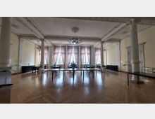 Ein großer, historischer Saal mit hohen Decken und verzierten Stuckelementen. Der Raum hat helle Wände, mehrere hohe Fenster mit rosa Vorhängen, Holzboden und Säulen, sowie Tische und Stühle, die gleichmäßig angeordnet sind.